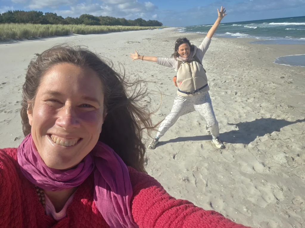 Ein Porträtfoto von Satyadevi und Yogita an der Ostsee auf der Insel Darß. Es ist sonniges und windiges Wetter. Sie lachen in die Kamera.